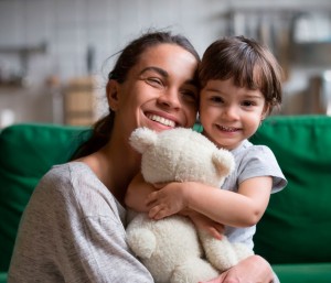 Mother hugging small child who is holding a teddy bear. Cute smiles on their faces