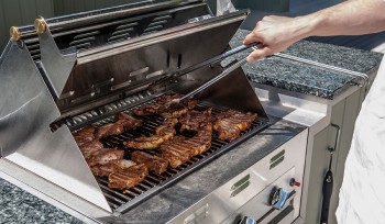 Hand holding tongs over large outdoor grill lined with meat cooking